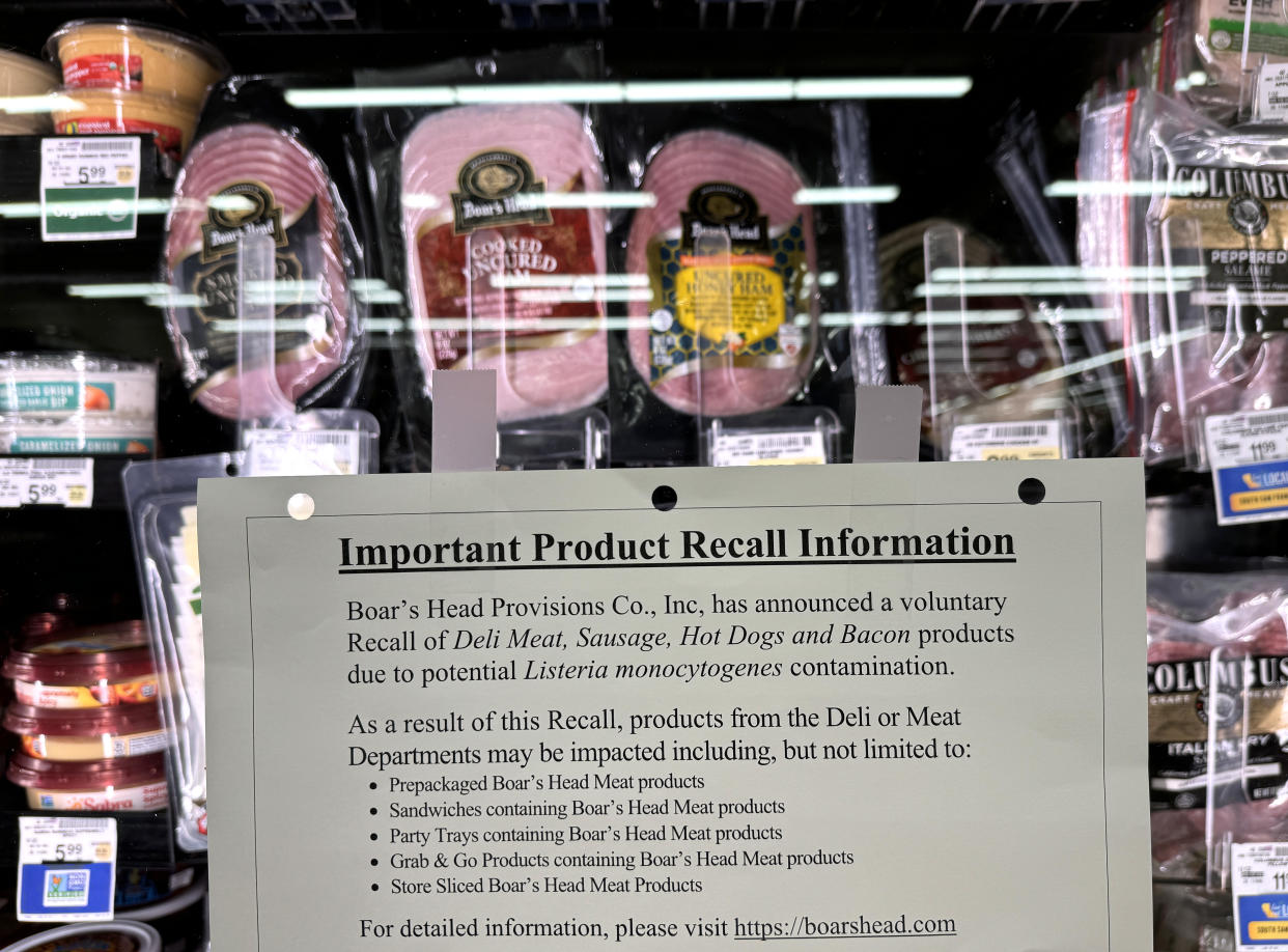 A product recall notice is attached to a grocer's refrigerated food case in front of packaged Boar's Head brand deli meats.