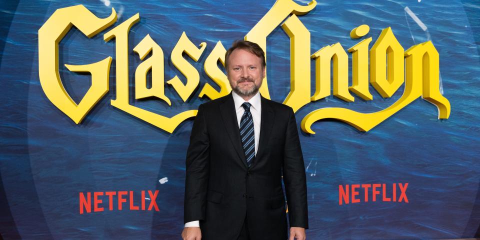 Rian Johnson in a suit and tie in front of a Glass Onion step and repeat