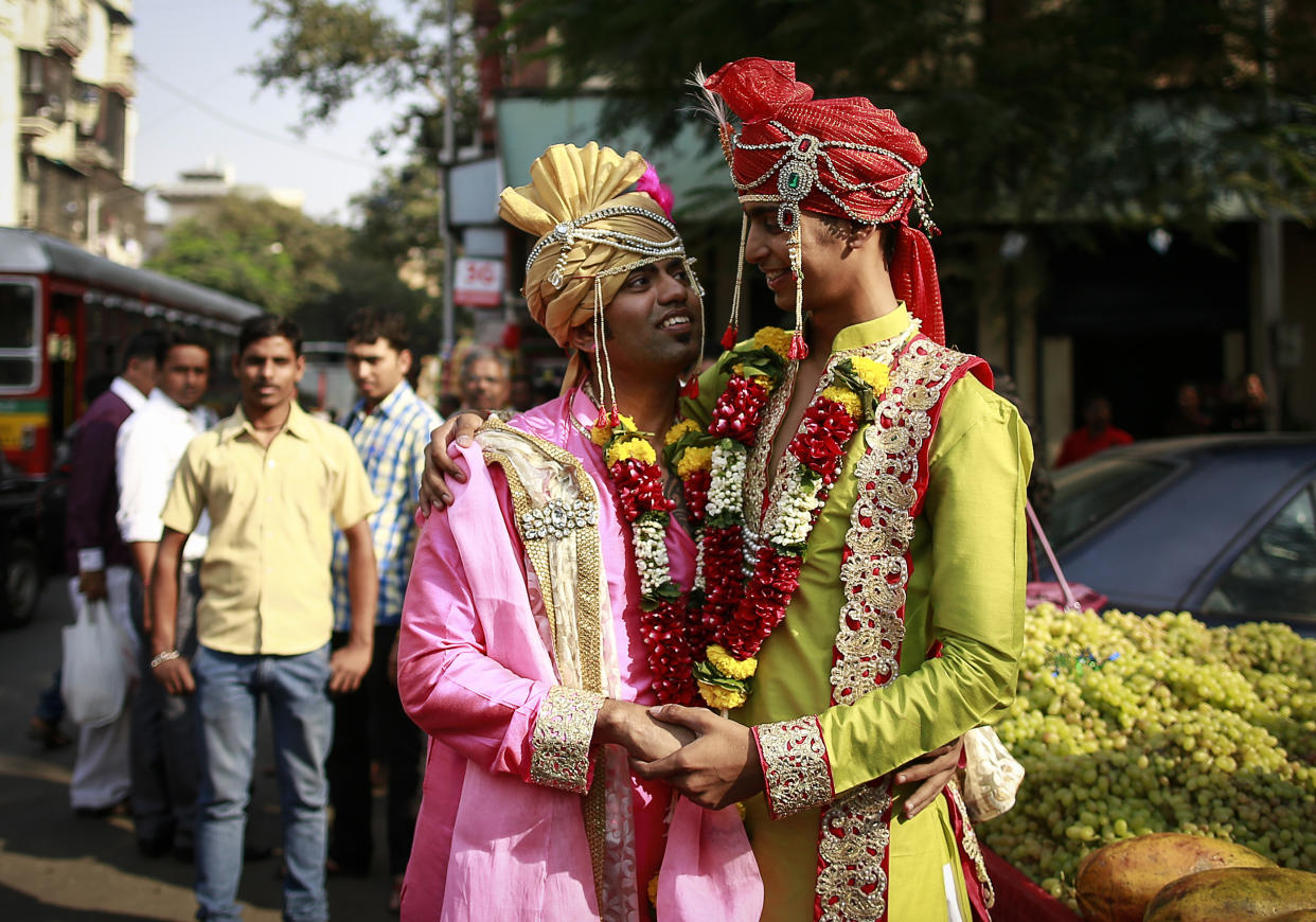 Two people in fancy attire embrace and hold hands on a street near onlookers.