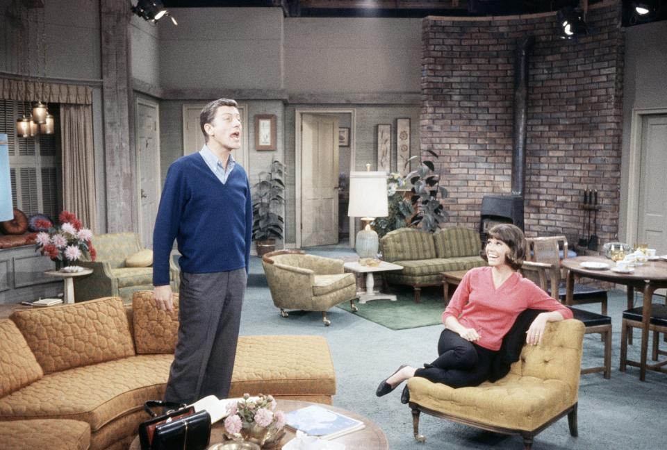 Dick Van Dyke and Mary Tyler Moore in a living room set, with Dick standing and Mary seated on a couch, both smiling warmly