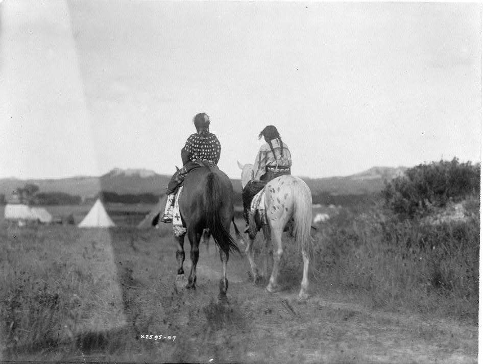 Two daughters of a chief on horseback, riding away from camera toward tents in background.