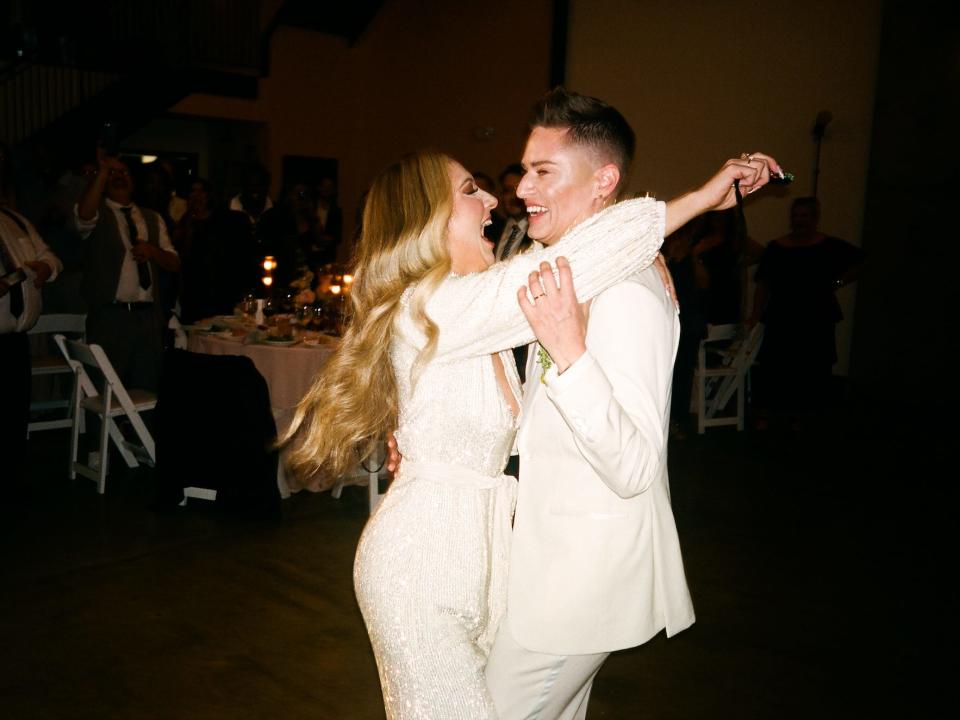 A bride in a white long sleeve dress and a bride in a white suit hug.