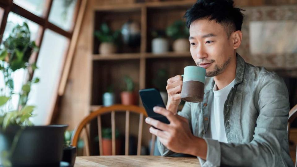 Young Asian man drinking coffee at home and looking at his phone