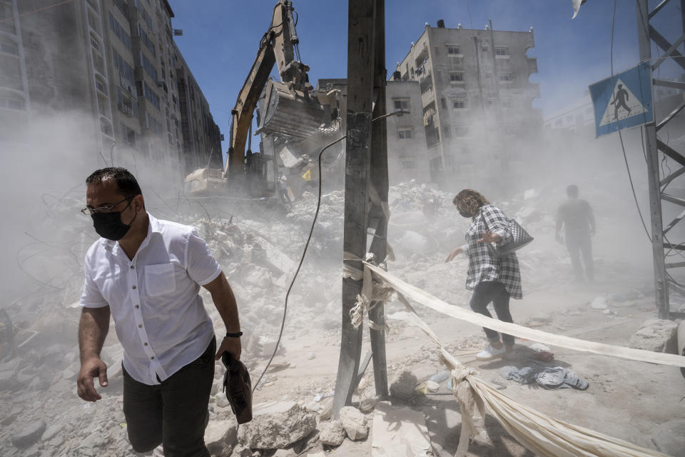 Pedestrians pass through clouds of dust as heavy construction equipment is used to sift through rubble to uncover valuables before it is transported away from the scene of a building destroyed in an airstrike prior to a cease-fire that halted an 11-day war between Gaza's Hamas rulers and Israel, Thursday, May 27, 2021, in Gaza City. (AP Photo/John Minchillo)