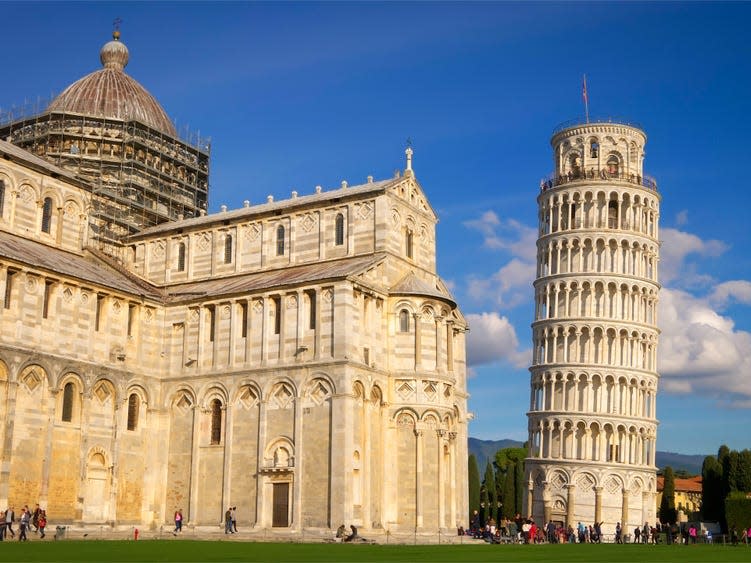 The Leaning Tower of Pisa and Cathedral against a blue sky. There are tourists walking around near both buildings.