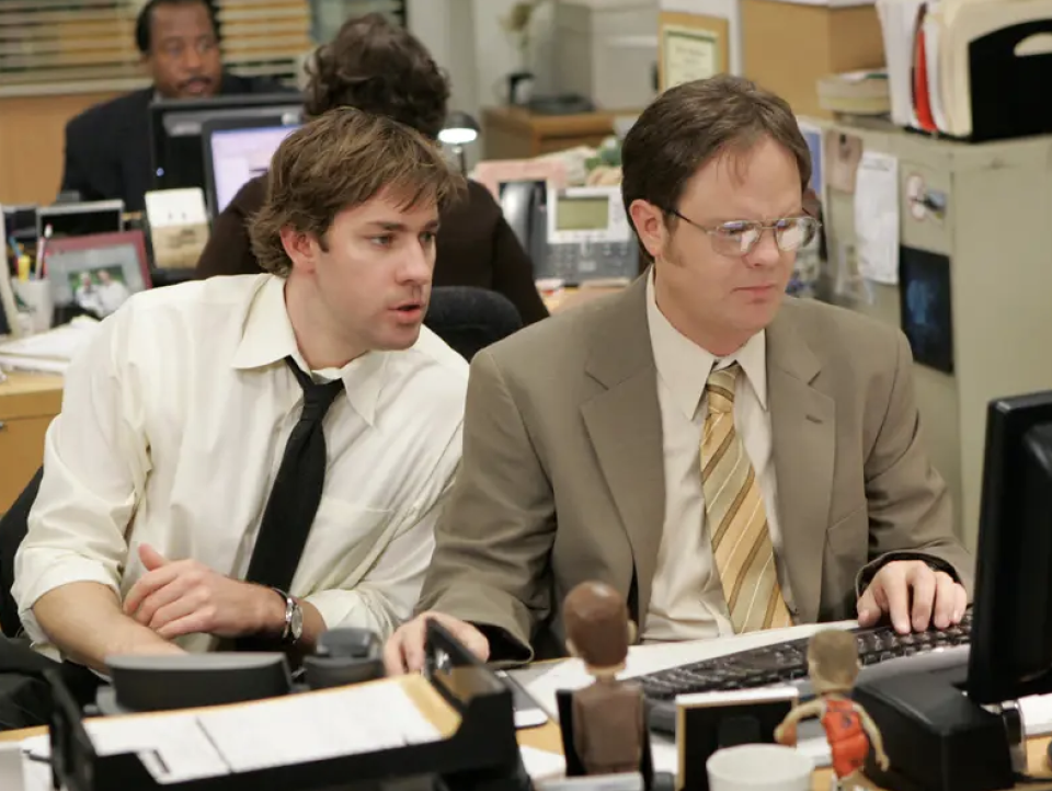 Two male characters from The Office, Jim and Dwight, seated at desks, appear to be working and discussing