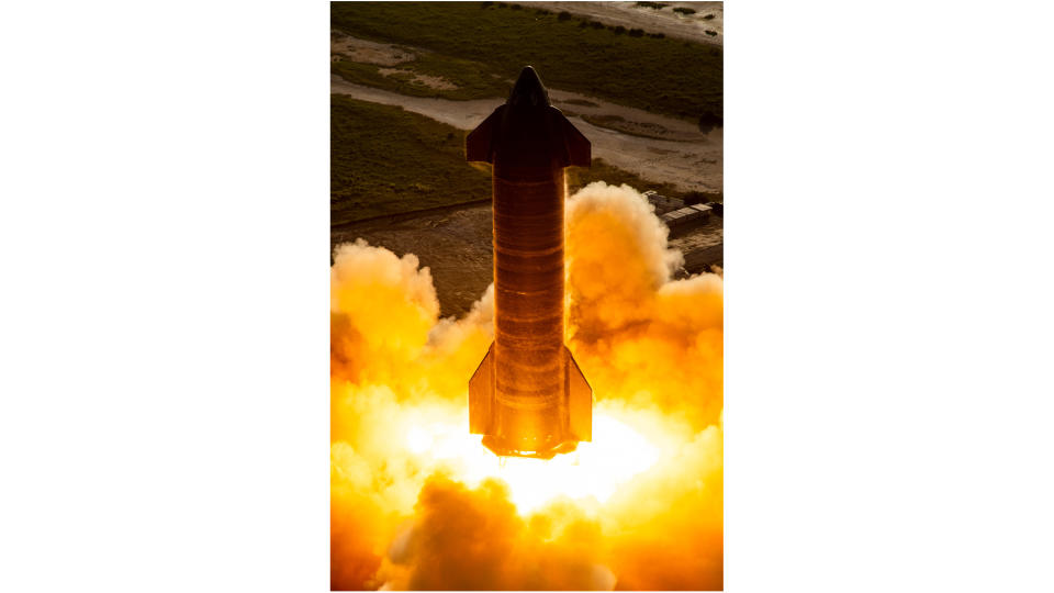 side-on view of shiny silver starship vehicle conducting an engine test, with orange flames and dust billowing around it