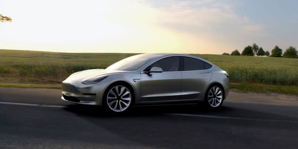 A silver Tesla Model 3 parked on a road, with a green field in the background