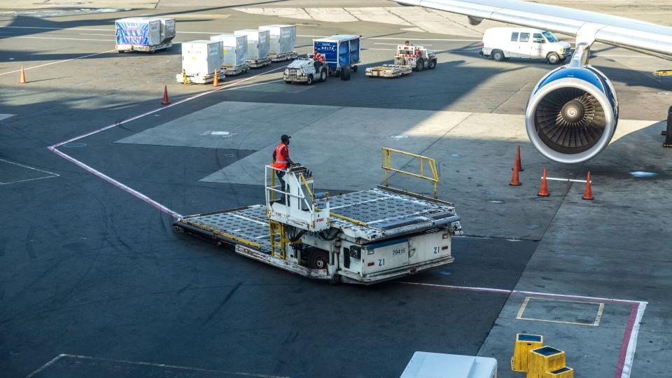 A worker maneuvers a cargo dolly near an aircraft with shipping containers lined up on the tarmac behind.