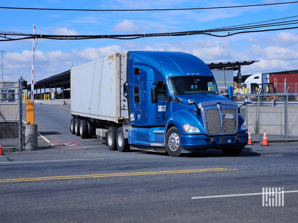 Image shows a blue semi hauling a refrigerated container out of a terminal.