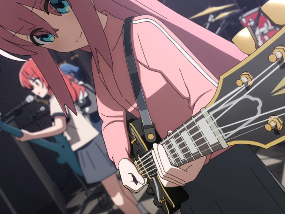 an animated girl with pink hair staring down at the neck of a black guitar, which she's playing. there' sa girl with red hair visible behind her, singing and holding a guitar