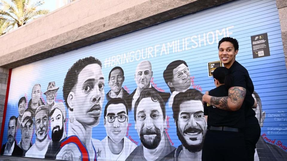 brittney griner hugs someone on her left and looks over her shoulder, she is smiling and both people wear all black outfits, they stand next to a painted mural of 15 people and the slogan bring our families home