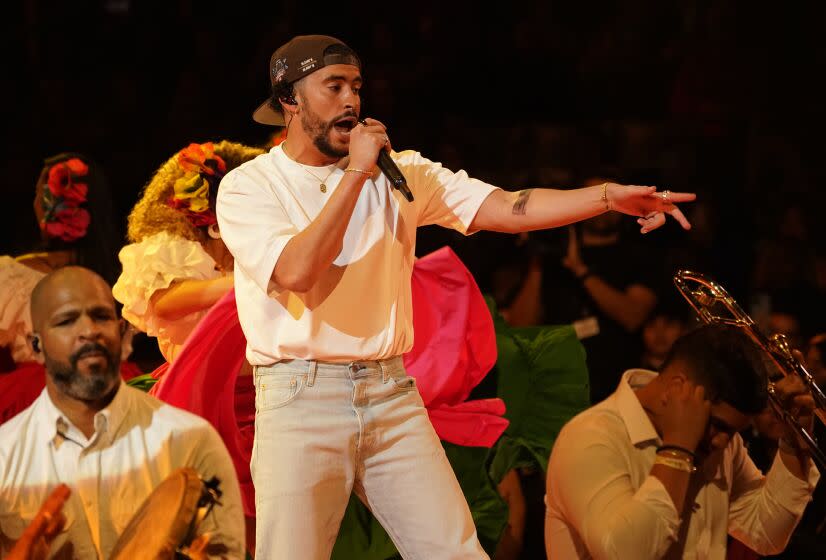 A man in a white T-shirt, jeans and a backward hat singing into a microphone amid a crowd of people with instruments