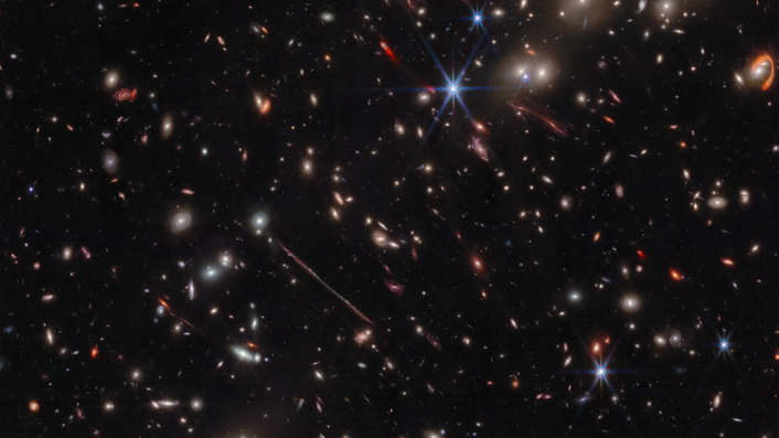  Various warped galaxies are seen as elongated lines, loops, arcs and smudges against the background of space. The JWST's iconic diffraction spikes are seen toward the center-right of the image. 