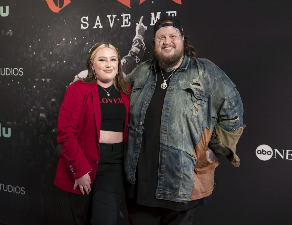 bailee deford and jelly roll smile and stand together for a photo, she wears a red jacket over a black crop top and black pants, he wears a denim jacket over a black shirt and pants with a backwards baseball cap