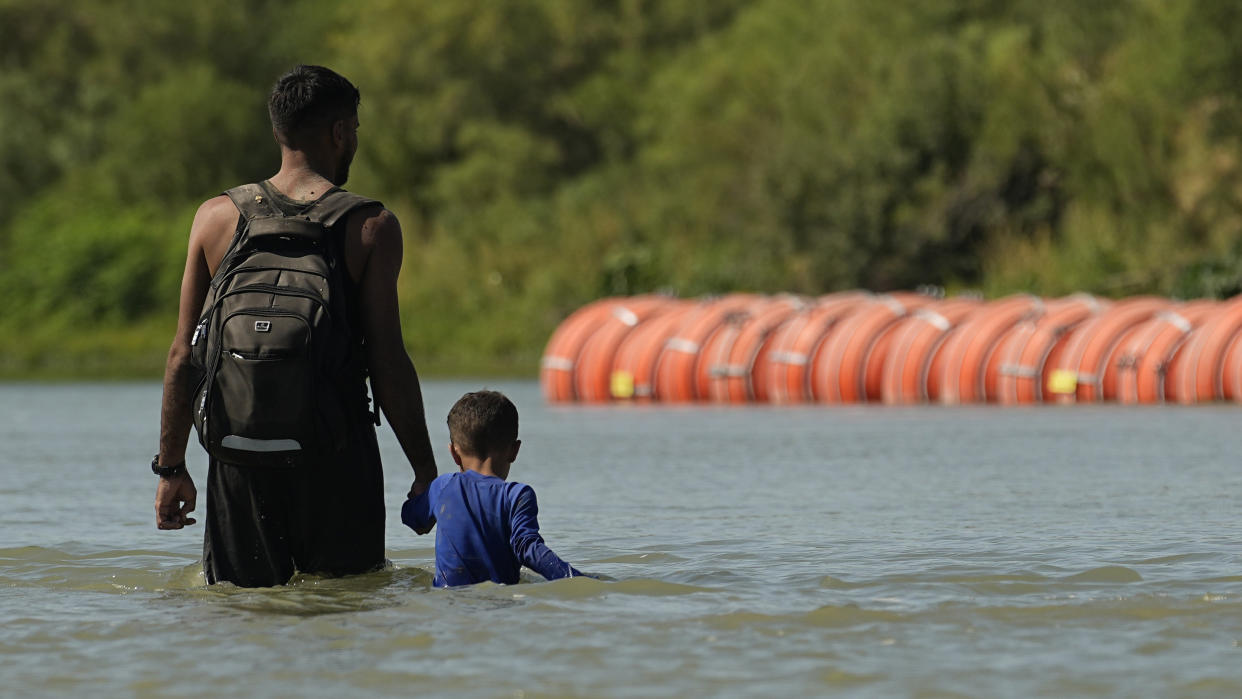 A man and child wade waist deep in the water near orange buoys.