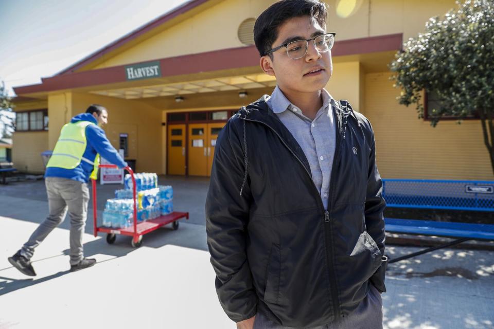 A young man stands in front of a building where another person pushes a cart with bottled water.