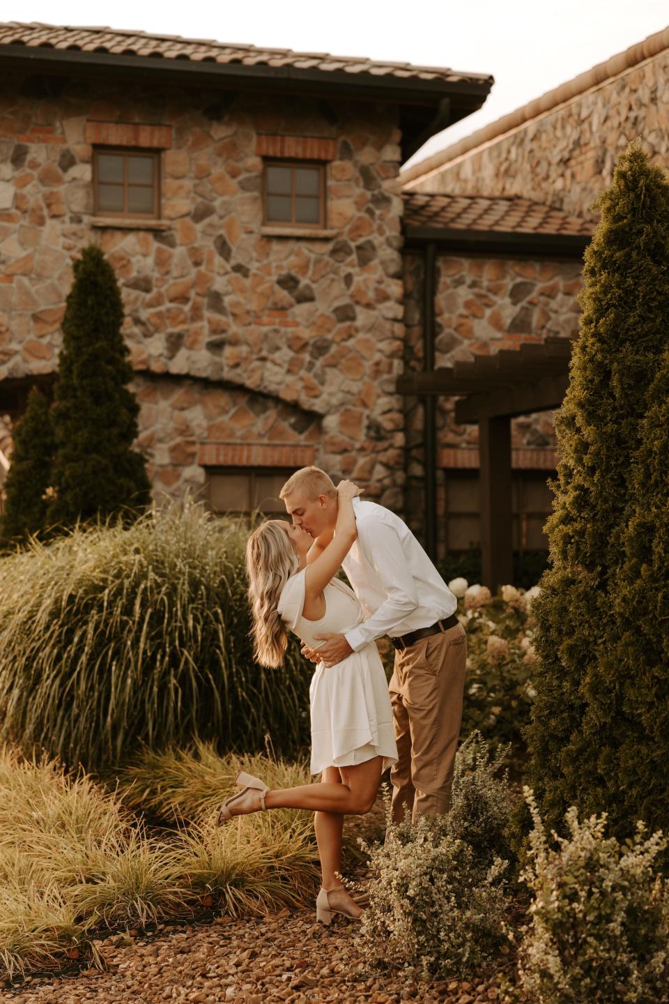 A woman in a white dress and a man in a white shirt and khakis kiss in front of a tuscan-looking building.