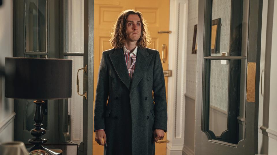 Jack Farthing as Selby in Rain Dogs standing in a hallway in an overcoat and striped shirt.