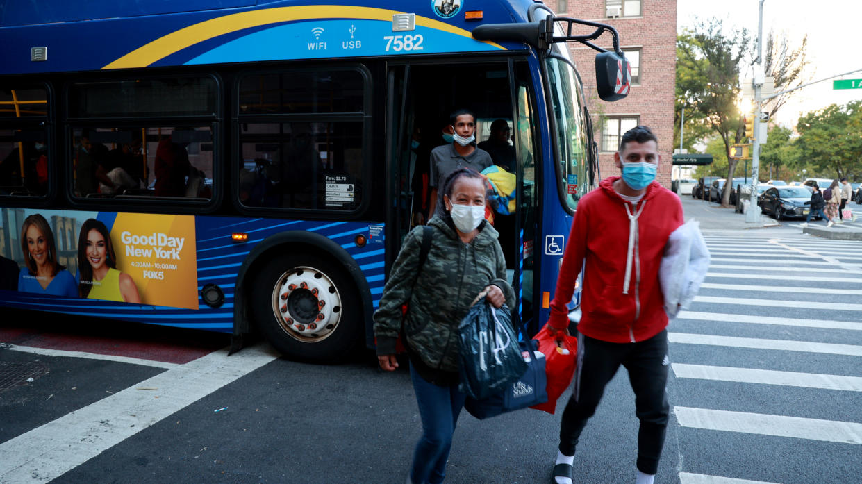 Three migrants wearing masks and carrying shopping bags of belongings get off a blue bus.