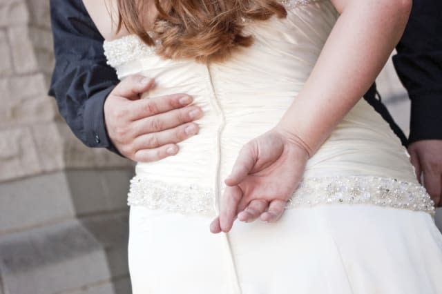 A bride stands with her back toward the camera, with her fingers crossed.