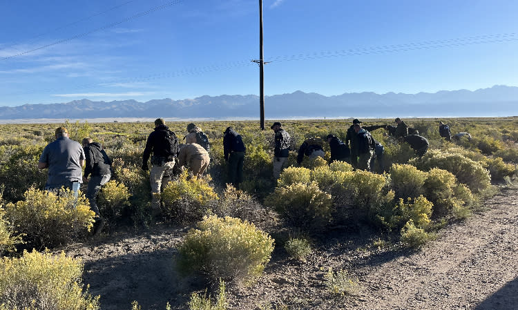 Law enforcement investigators in a field of brush with the Colorado Rocky Mountains in the background