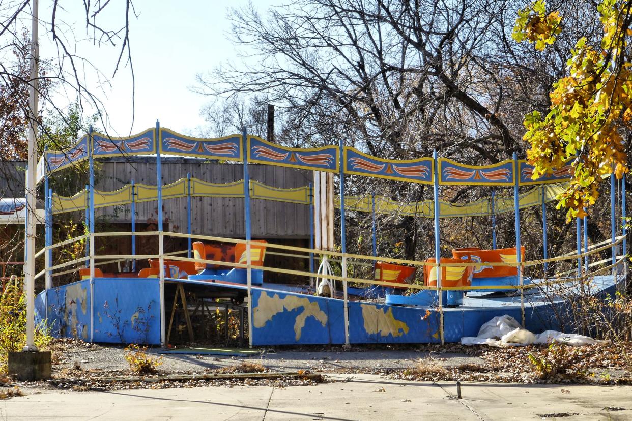 Abandoned Joyland Amusement Park, Wichita, Kansas, broken tilt-a-whirl ride fallen apart, sidewalk in the foreground, one tree with colored leaves on the foreground right, surrounded by bare trees, on a sunny day