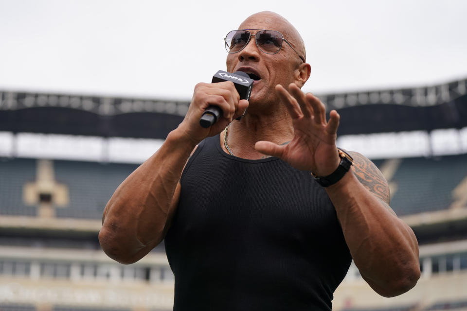 Dwayne Johnson speaks into a microphone, wearing a black tank top at an outdoor event