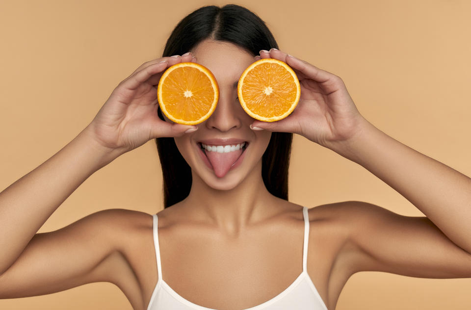 Portrait of Asian girl with shining clean skin of face holding orange halves in white underwear isolated on beige background. Vitamin C cosmetics concept