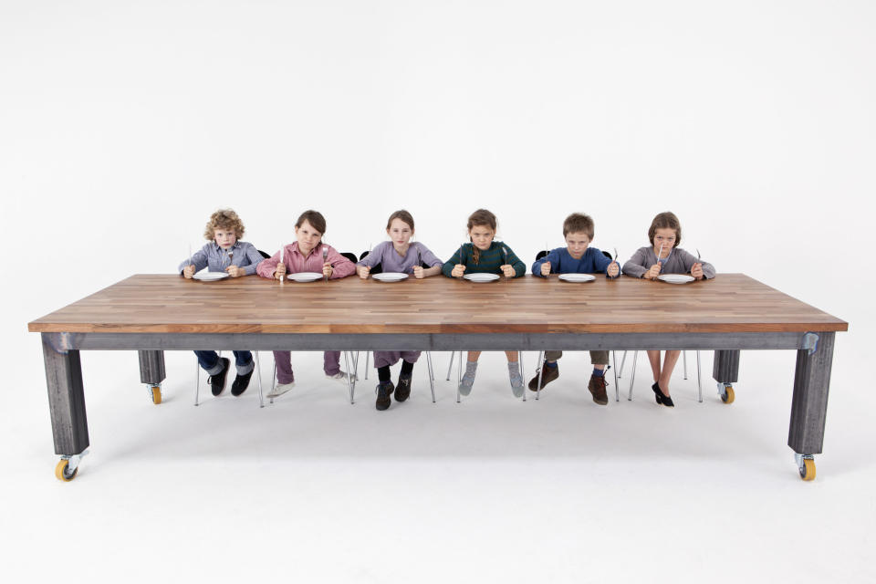 Seven children sit at a long wooden table with empty plates, looking serious and expectant