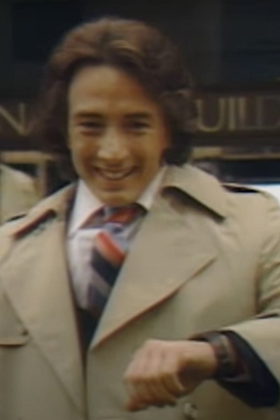 A young Martin Short, in a beige trench coat smiling at the camera and holding out their arm