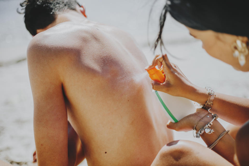 A person sprays sunscreen onto another individual's bare back on a sunny beach day, emphasizing proper skin protection