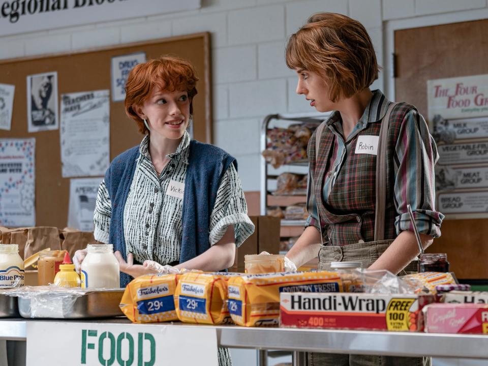 vickie and robin in stranger things, standing at a set-up folded table in a gym making peanut butter and jelly sandwiches and looking towards each other