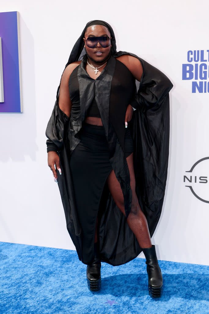 Saucy Santana  on a blue carpet wearing a black avant-garde outfit with a hooded cape, large sunglasses, and black platform boots