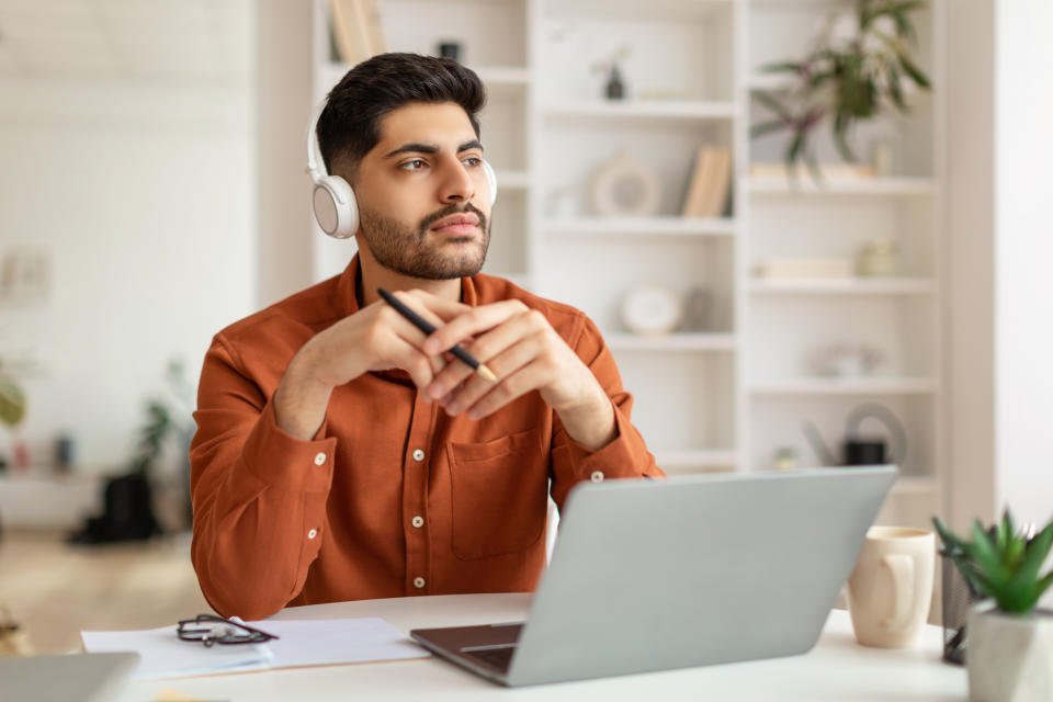 Portrait Of Pensive Focused Middle Eastern Male Manager In Wireless Headset Working On Laptop Computer In Home Office, Serious Guy Sitting At Desk And Using Pc, Looking Away Thinking About Job