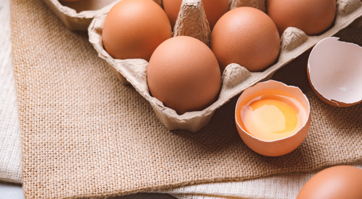 A carton of eggs is placed on top a burlap bag. One egg is cracked open. Why is the cost of eggs rising?