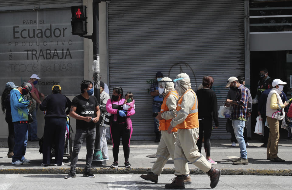 Workers dressed in protective from head to toe walk past pedestrians wearing protective face masks as a mandatory measure to help curb the spread of the new coronavirus, walk past in downtown Quito, Ecuador, Wednesday, June 10, 2020. The city is returning to a new normality after relaxing a rigorous quarantine but amid a certain fear that COVID-19 infections may rise. (AP Photo/Dolores Ochoa)
