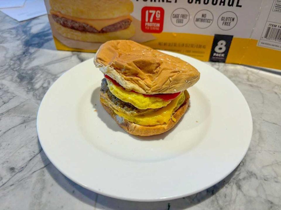 An egg'wich, with two egg patties, a sausage patty, and cheese, in between a hamburger bun on a white plate with yellow and white box behind it