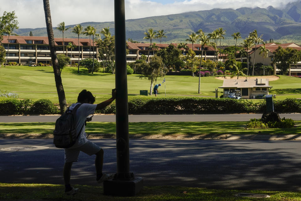 A boy waits for a shuttle as a man golfs at the Ka'anapali Golf Courses, Wednesday, Dec. 6, 2023, in the beach resort community of Kaanapali in Lahaina, Hawaii. Residents and survivors still dealing with the aftermath of the August wildfires in Lahaina have mixed feelings as tourists begin to return to the west side of Maui. (AP Photo/Lindsey Wasson)