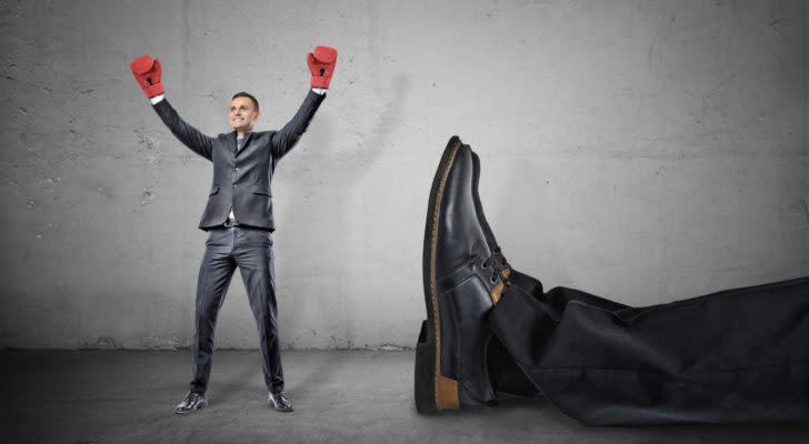 a tiny man with boxing gloves standing next to the feet of a giant man whom he presumably knocked out