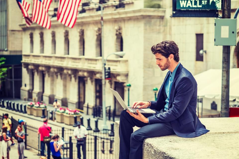 A man sitting on a ledge looking at laptop in front of a street sign reading Wall St.