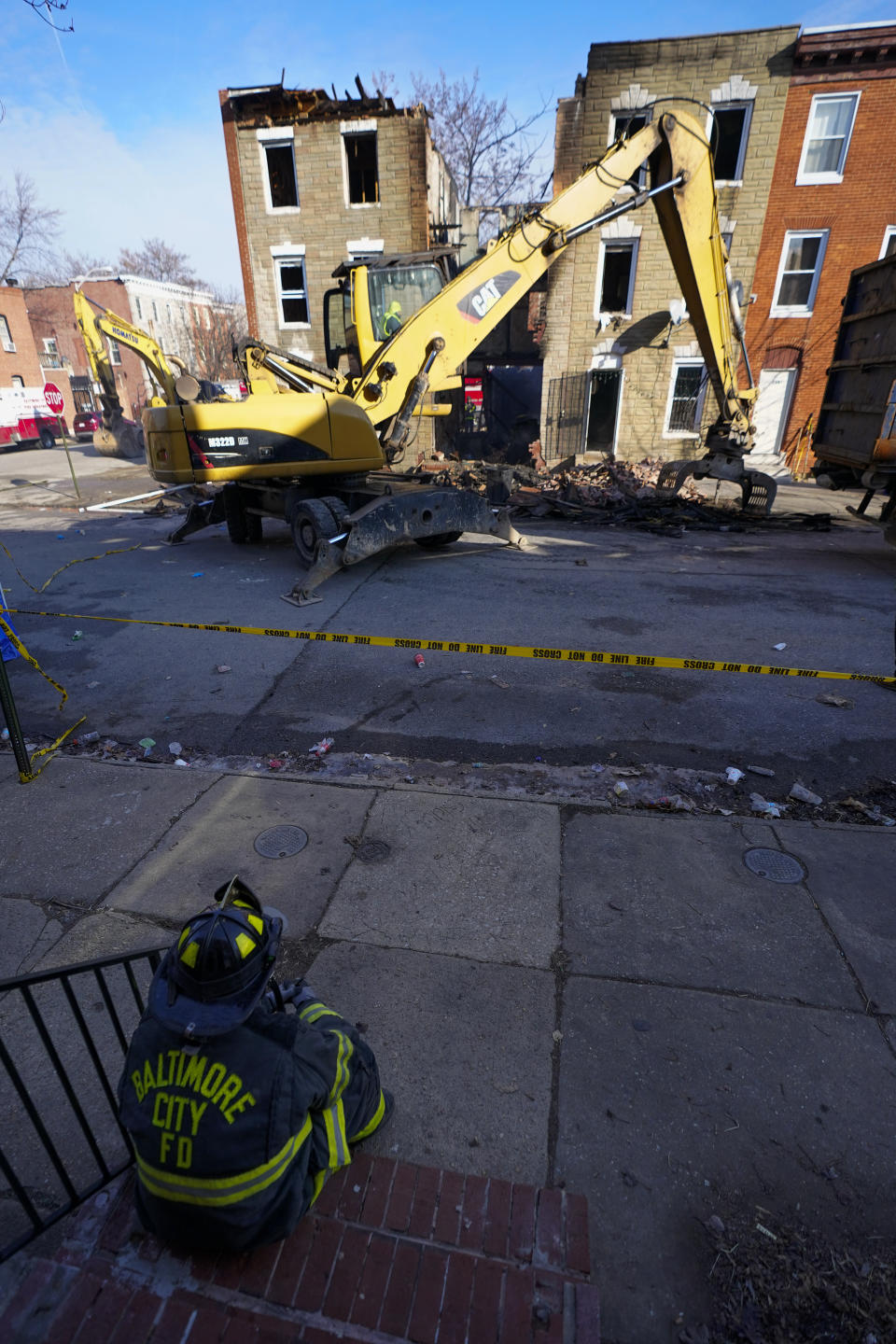 A firefighter sits on the stoop of a home as he watches an excavator pull debris off a building during efforts to retrieve the body of a deceased firefighter caught in the building's collapse while battling a two-alarm fire at the vacant row home, Monday, Jan. 24, 2022, in Baltimore. Officials said several firefighters died during the blaze. (AP Photo/Julio Cortez)