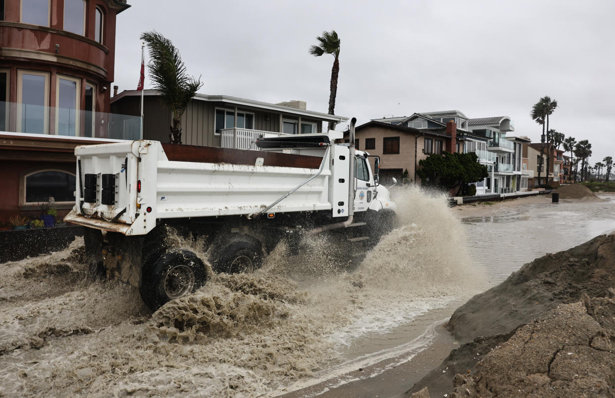 A worker drives a truck through standing water while creating sand berms to protect beachfront homes from flooding.