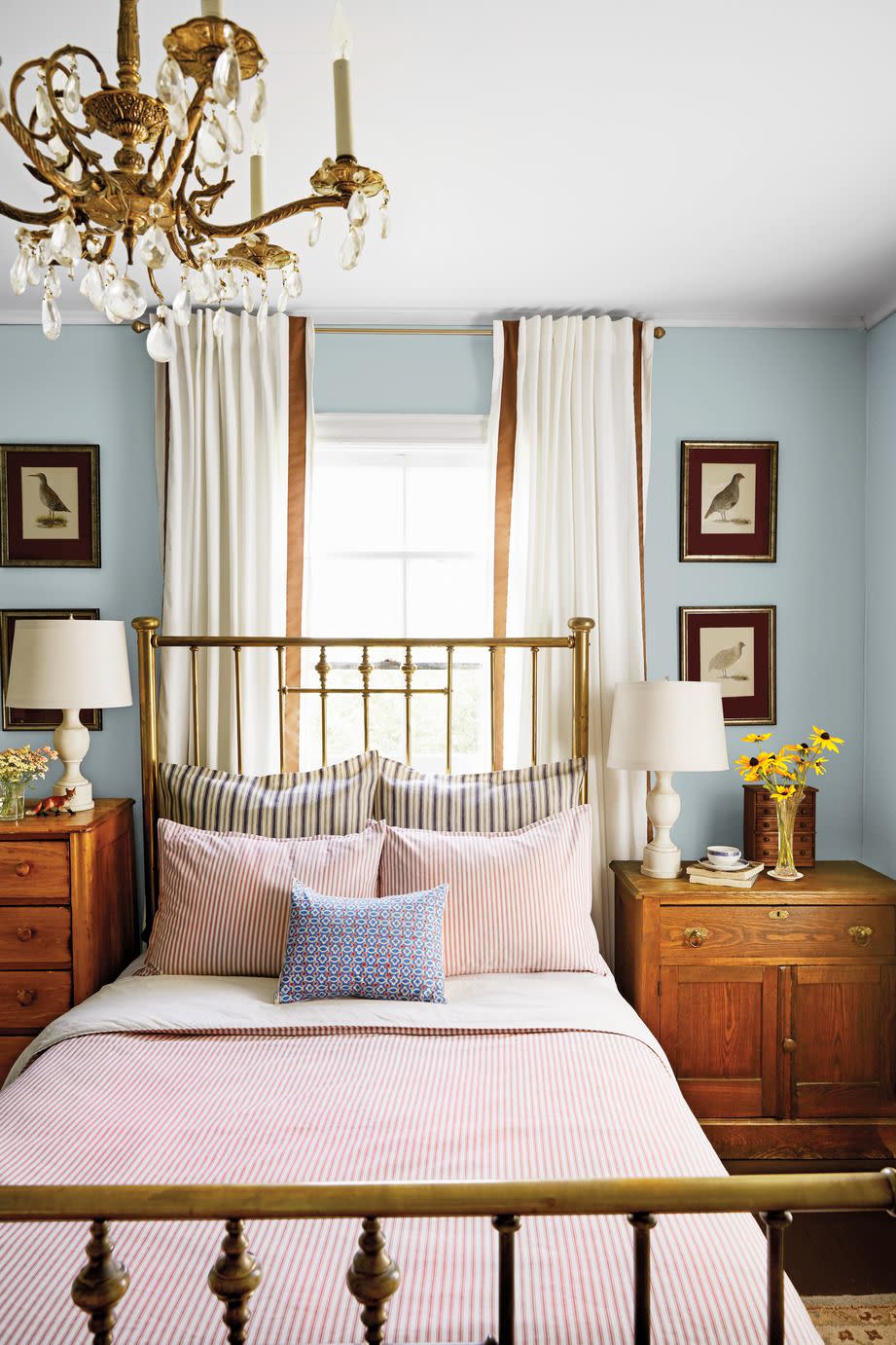 the homes red and blue palette takes a moodier turn in this bedroom with an antique brass bed with a dramatic 54 inch tall headboard that adds to the sophisticated look and mismatched dressers and 99 cent bird prints flank each side of the piece