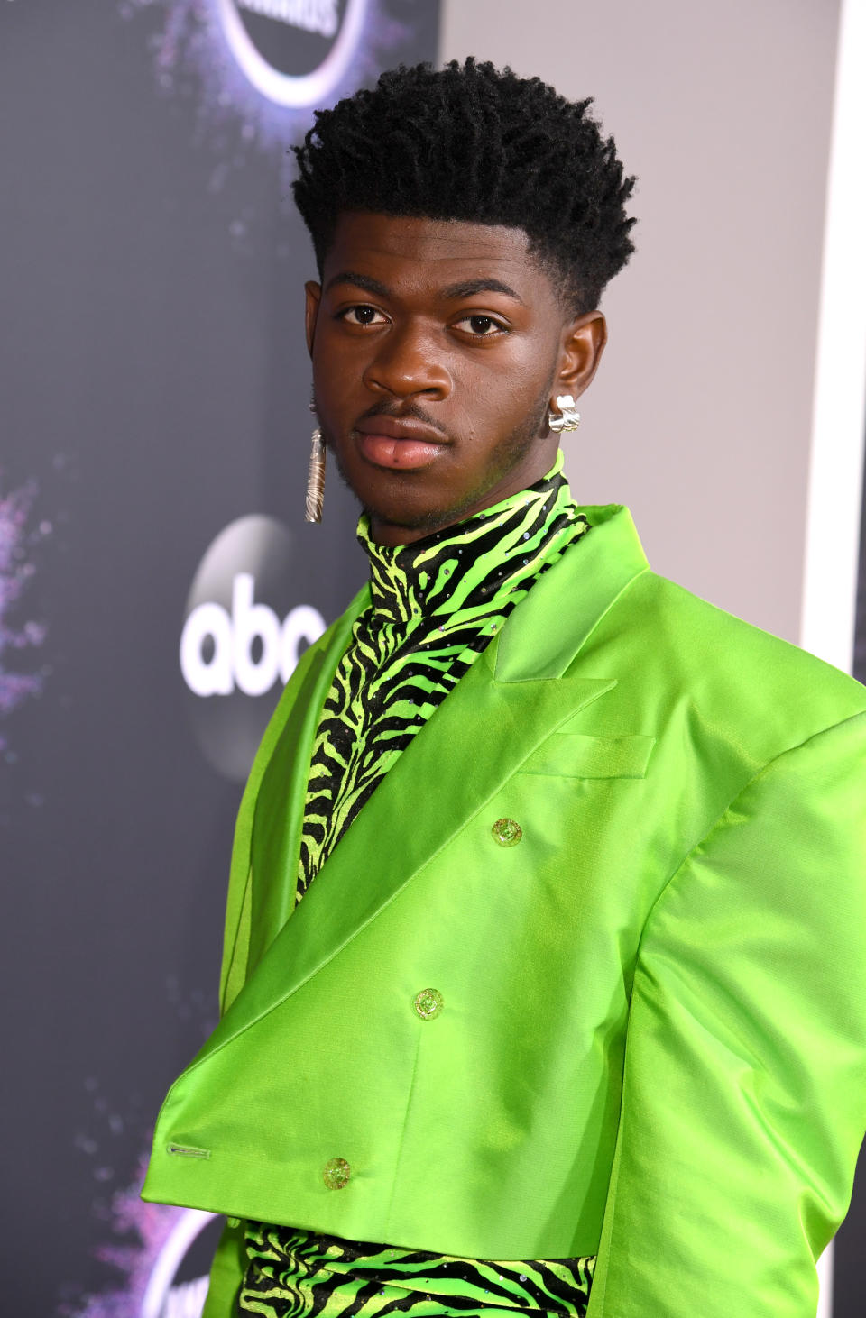 Lil' Nas X at a red carpet event, wearing a neon suit with a zebra print turtleneck and a dangling earring