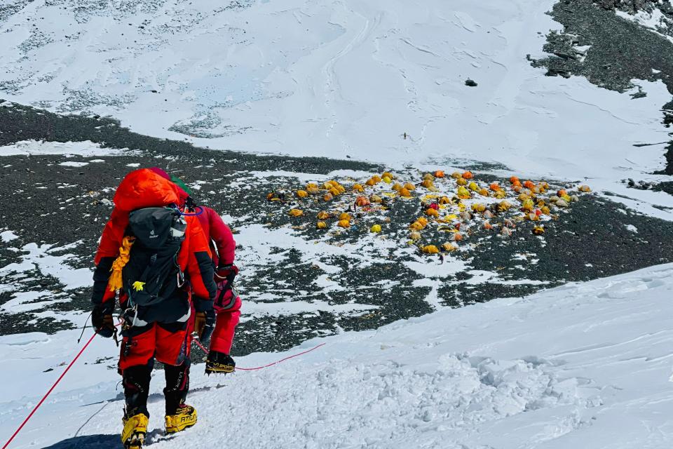 Two mountaineers in red coats standing at the edge of a large drop with a snow-covered expanse in front of them.