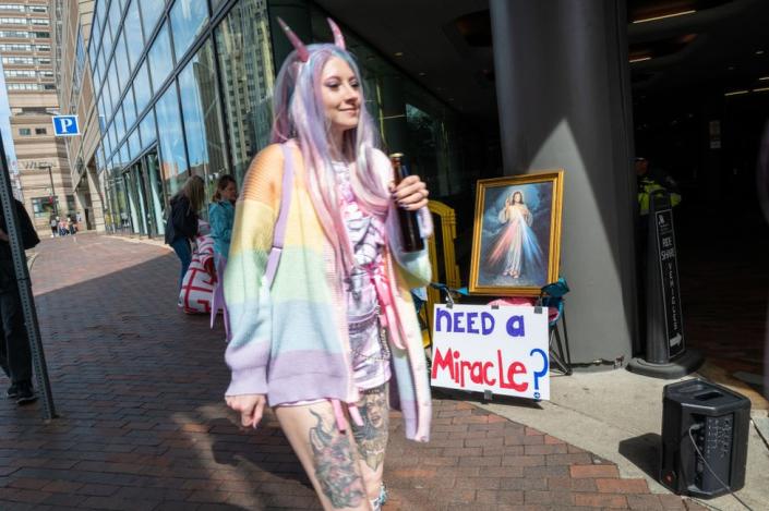 Outside the Marriott Copley hotel, a woman in a long pink and blue wig - wearing pink horns and a rainbow cardigan - smiles as she walks past a sign that reads: &quot;Need a miracle?&quot; and a picture of Jesus.