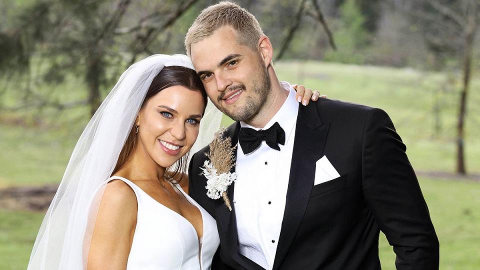 2021 MAFS couple Coco and Sam pose in a garden at their show wedding. A large veil covers her dark hair and she wears a gown with a plunging neckline. He is in a black suit with bow tie and has a short beard and moustache. 