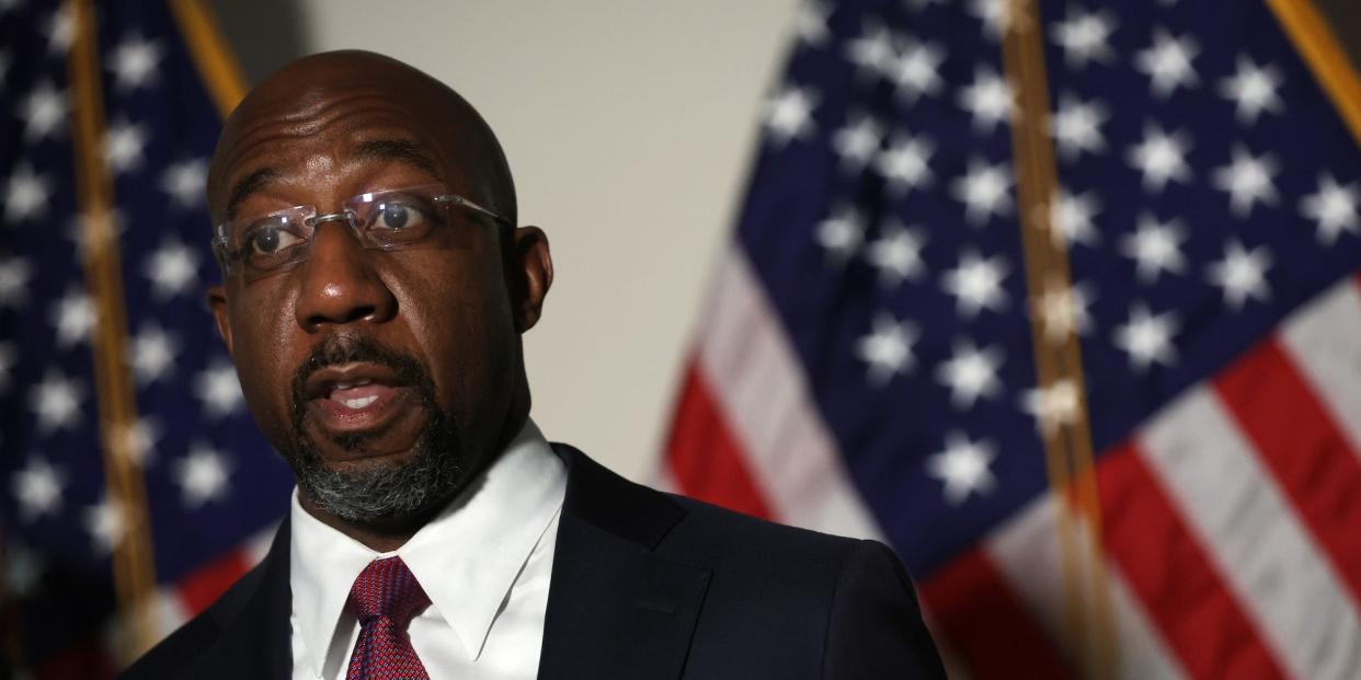 Suit-clad Democratic Sen. Raphael Warnock of Georgia speaks to reporters while standing against a backdrop of American flags at the US Capitol.