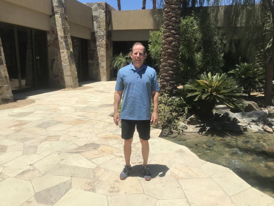 A man in a light-blue short-sleeved shirt and dark shorts stands in a landscaped courtyard